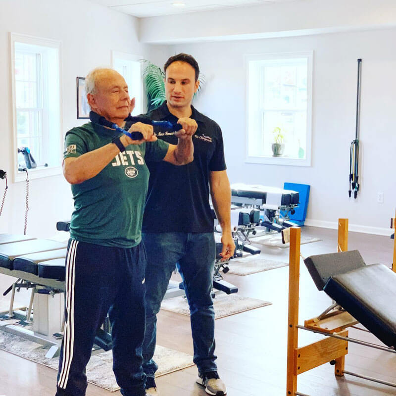Dr. Marco Ferrucci, a chiropractor, guiding an elderly patient with resistance band exercises at 'The Chiropractic Source' clinic.